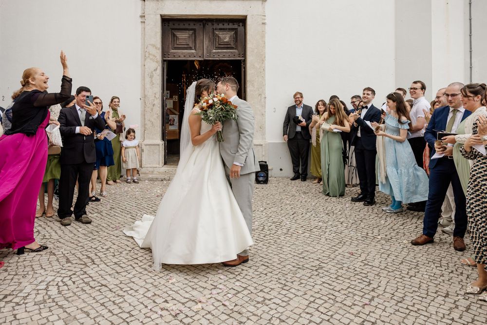 Bride and groom share a kiss outside a church surrounded by cheering wedding guests on a cobblestone courtyard.