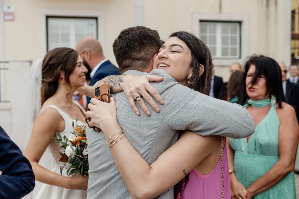 Guests sharing warm hugs and laughter at an outdoor wedding celebration, with bride holding a colorful bouquet nearby.