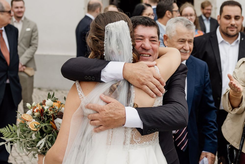 Bride in white gown and veil hugging guests while holding a floral bouquet at an outdoor wedding celebration.
