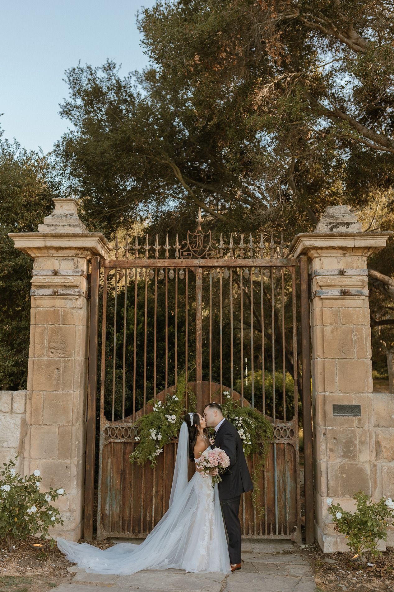 Bride and groom kissing before ornate iron garden gates adorned with florals at The Havens Country Club in Vista California.