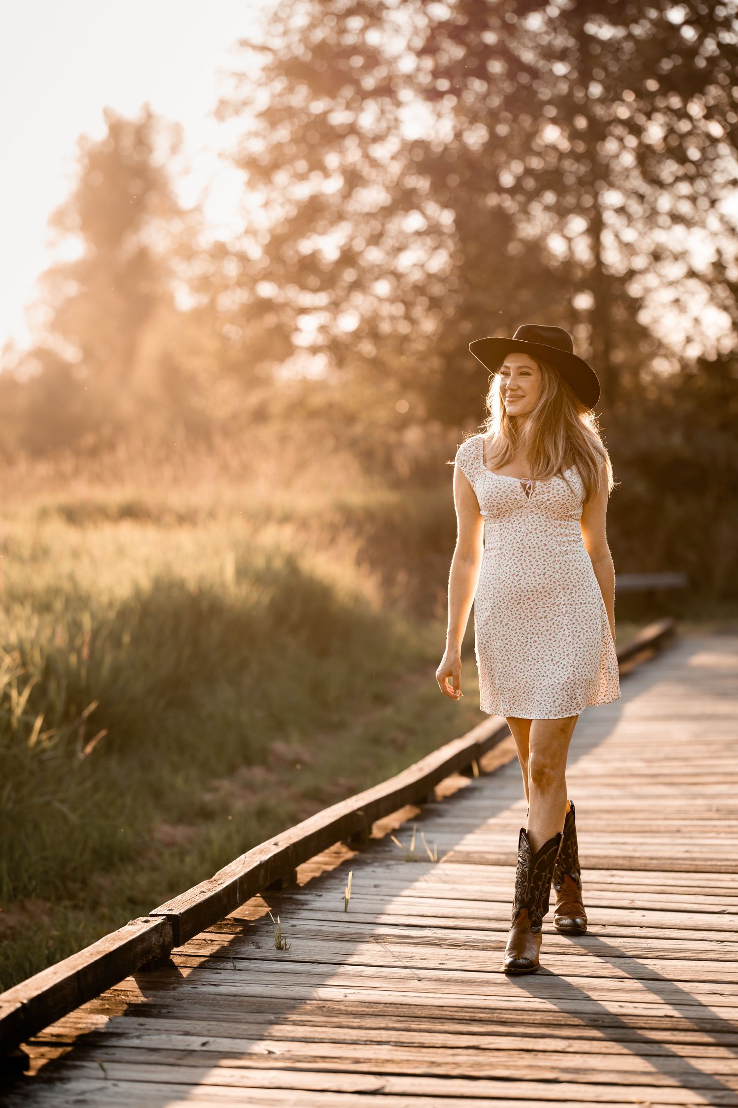 Woman in white dress, cowboy hat, and boots walks on wooden boardwalk at golden sunset.