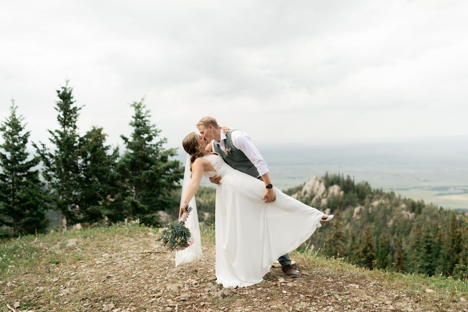A couple shares a romantic kiss during an elopementon a Montana mountaintop with evergreen trees and a misty valley vista in the background.