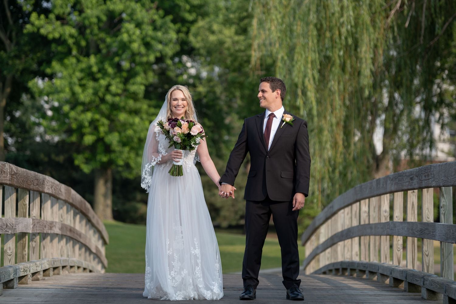 A bride and groom hold hands while standing on a wooden bridge with a weeping willow tree in the background.