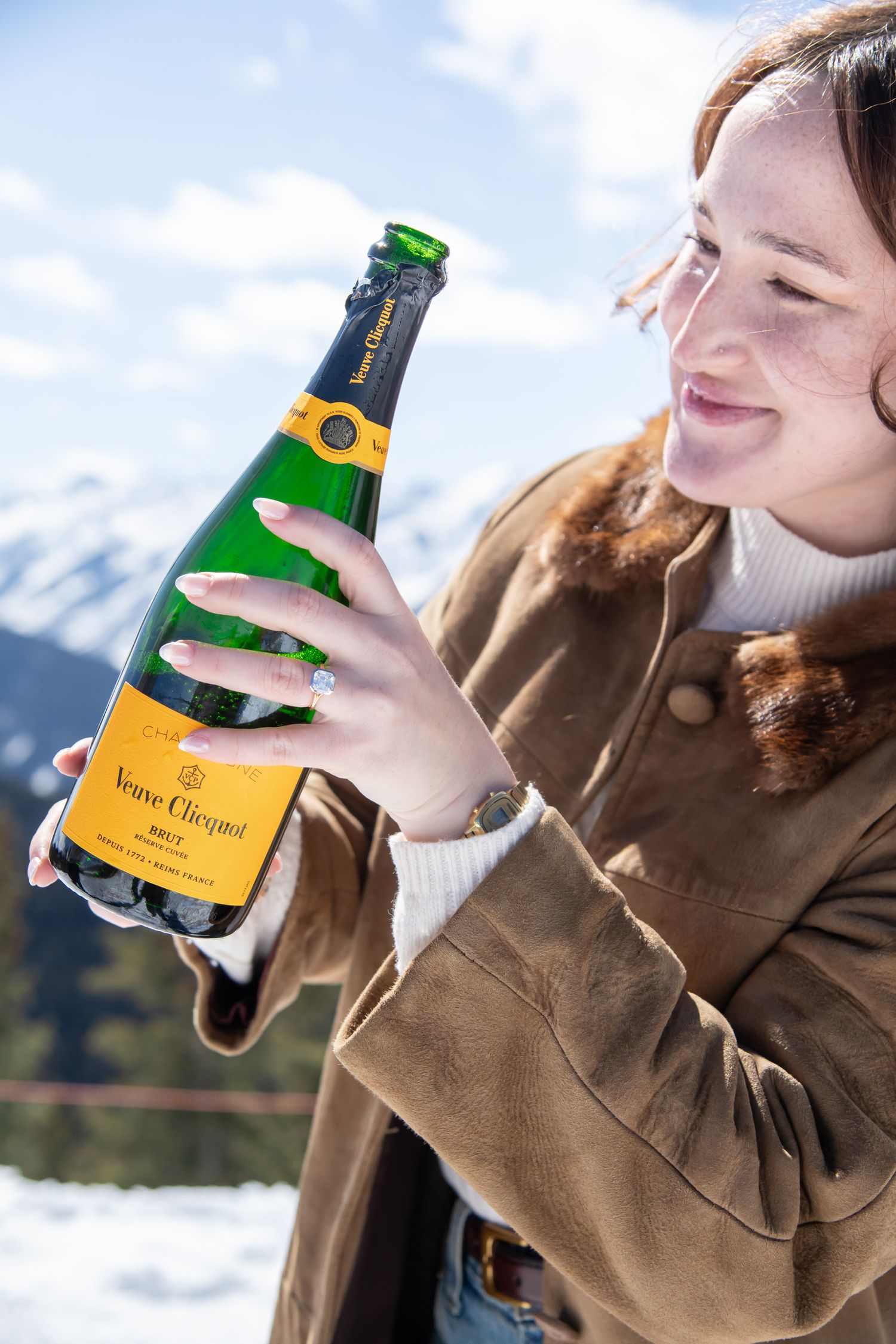 Person in brown coat drinking from a green champagne bottle with snowy mountains in background in Aspen Colorado engagement.