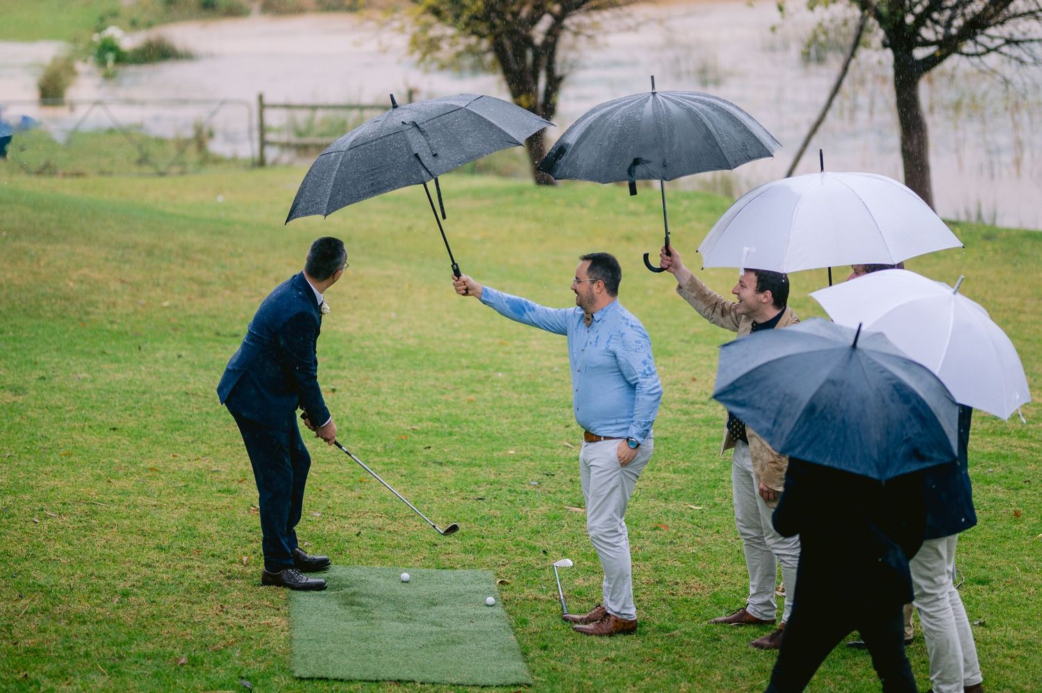 Men playing golf in the rain while holding black and white umbrellas on a lush green course.