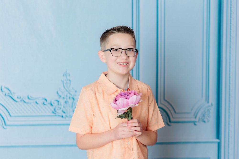 A person in a peach-colored shirt holds a pink peony flower against a light blue wall with decorative molding.