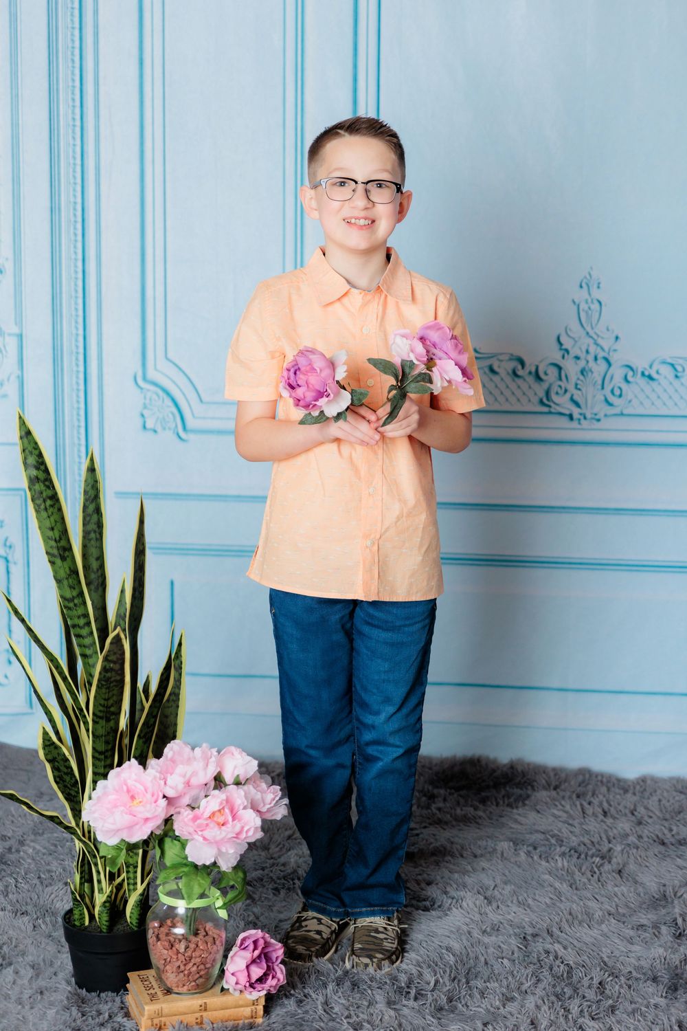 Person stands near pink flowers and potted plants against blue wall with ornate trim details.