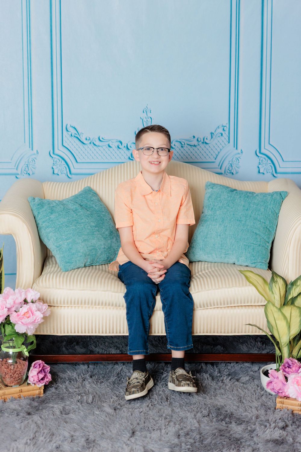 Person sits on cream colored couch with teal pillows next to decorative plants and pink flowers.