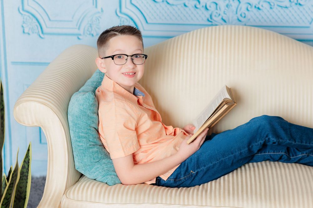 Reader in casual attire enjoys quiet time with book on classic upholstered sofa near potted plants.
