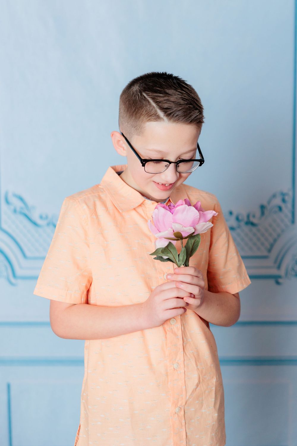 A person in an orange shirt smells a pink carnation flower against a blue wall background.