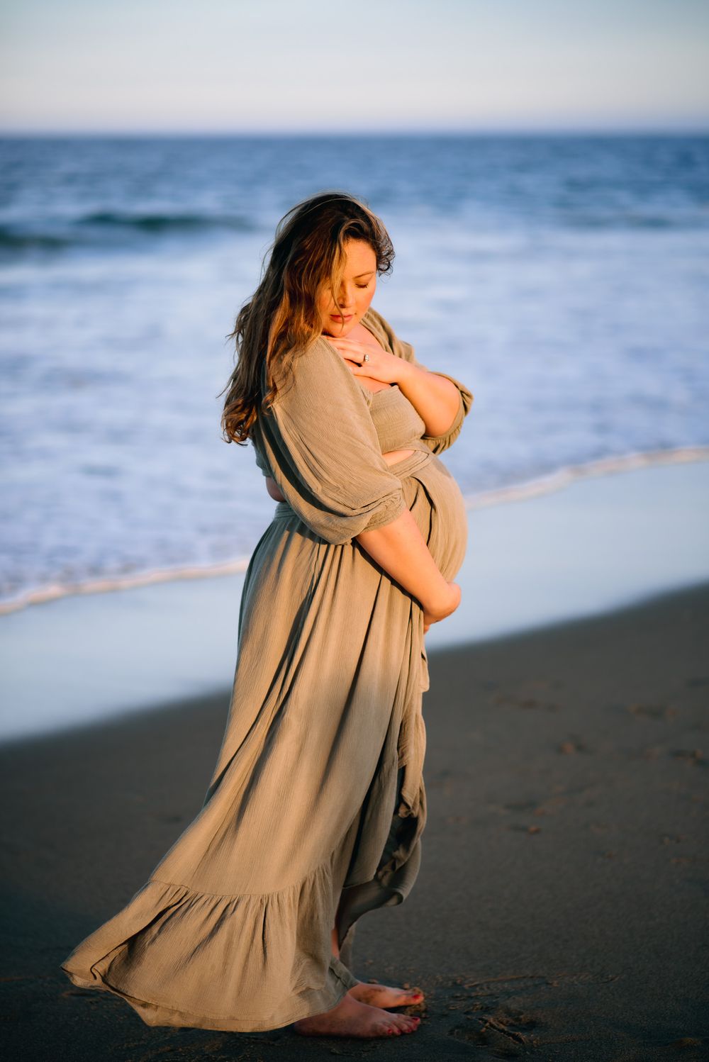 pregnant mother in flowing dress during golden light standing on the ocean beach.