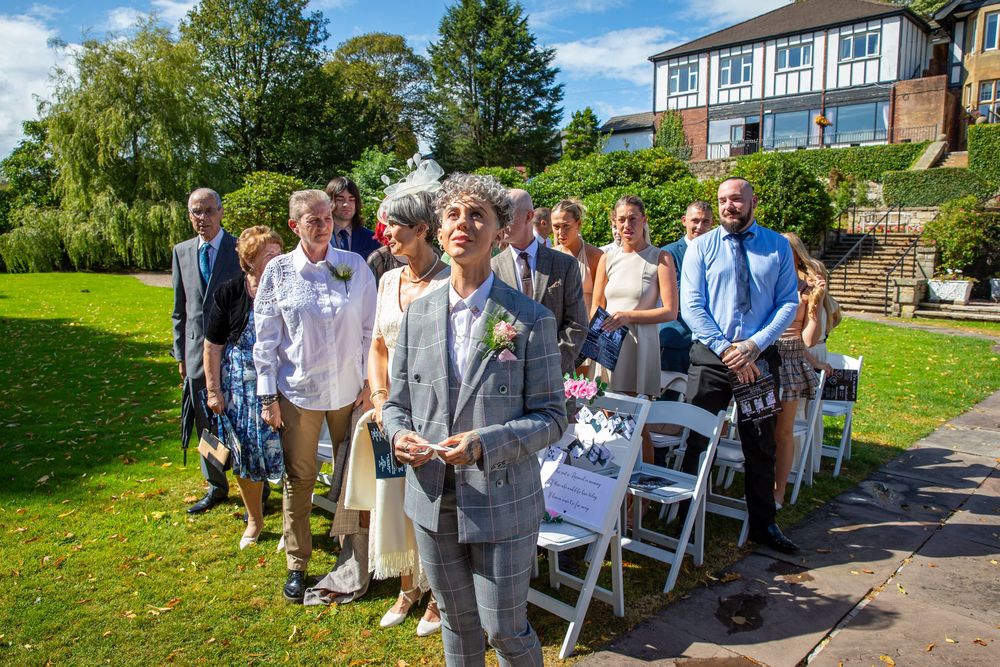 Wedding officiant addresses guests seated outdoors on white chairs at a garden wedding ceremony.