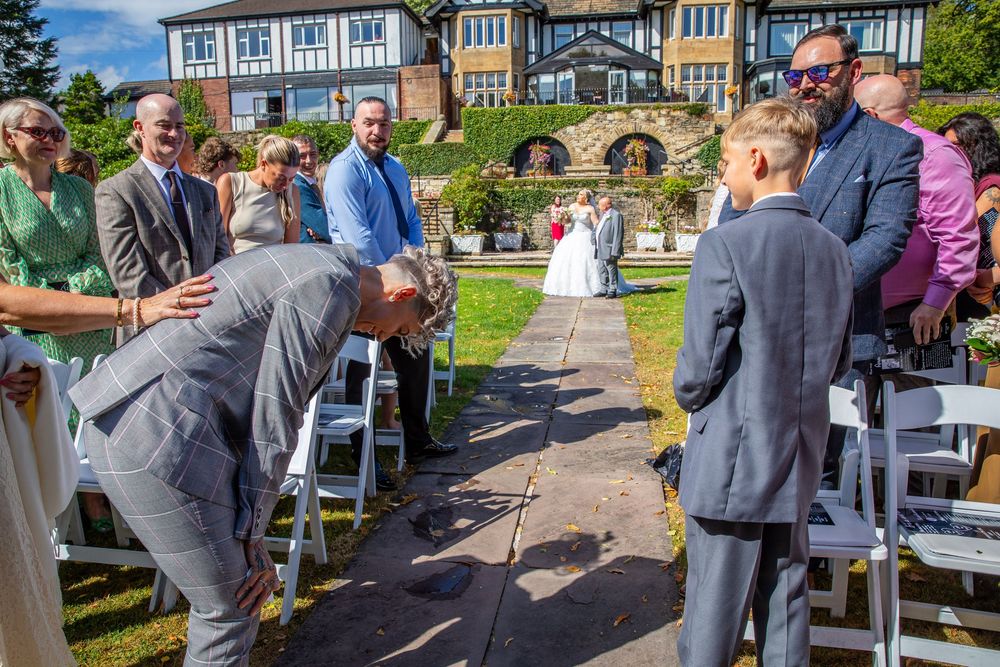 Groom bowing to young boy at outdoor wedding ceremony aisle with Tudor mansion backdrop.