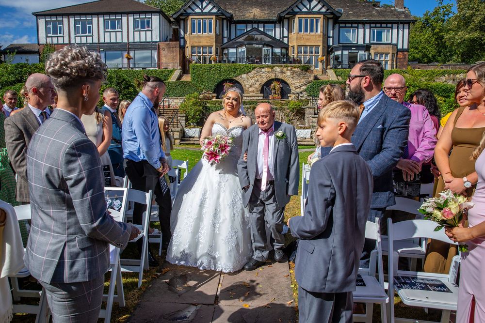 Bride walking down aisle with father at elegant outdoor wedding ceremony beside Tudor mansion.