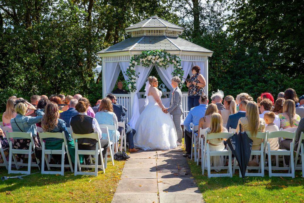 Couple exchanging vows at outdoor wedding ceremony beneath white gazebo surrounded by seated guests.