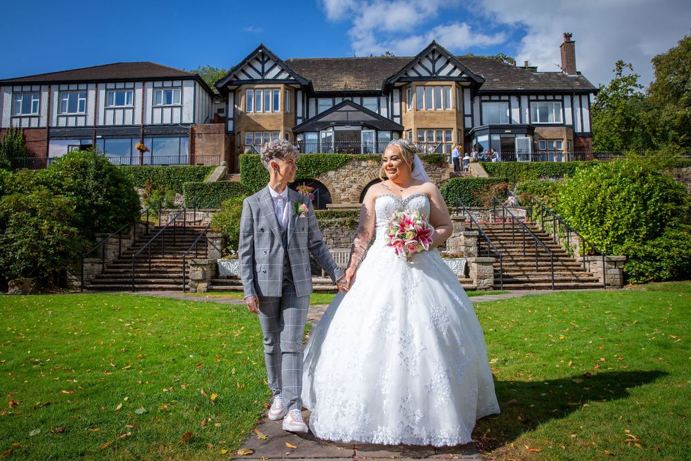 Newlywed couple posing in garden in front of grand Tudor-style manor on sunny wedding day.