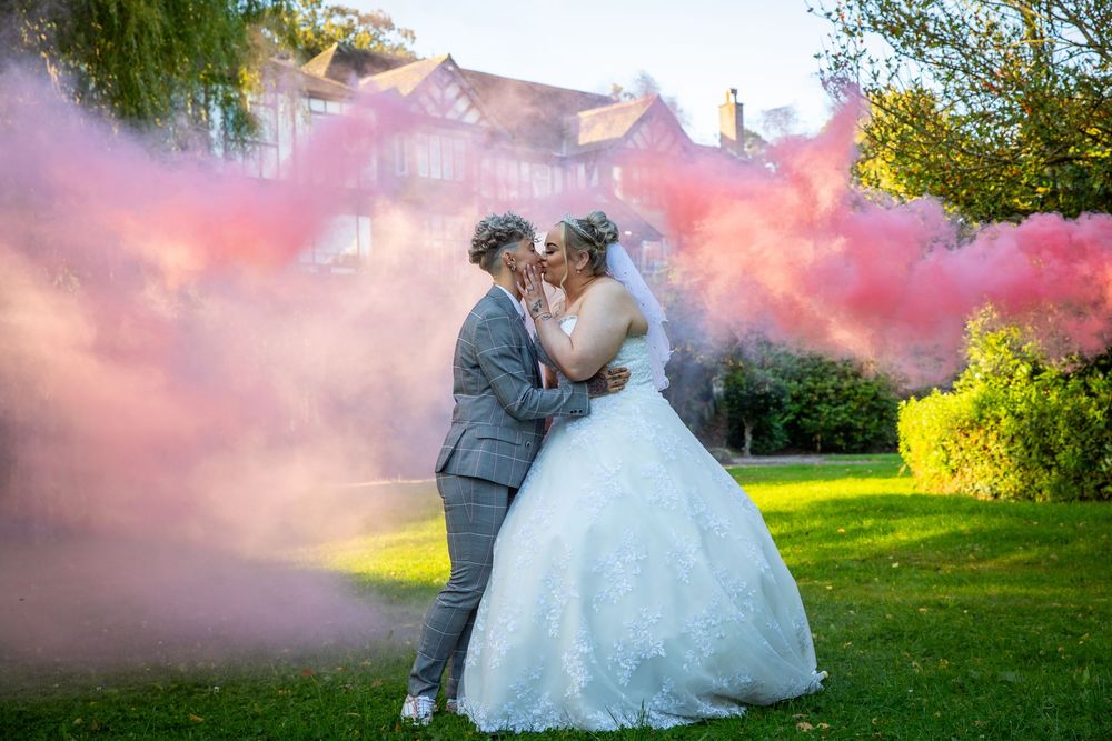 Newlywed couple kissing surrounded by vibrant pink smoke bombs in lush garden setting.