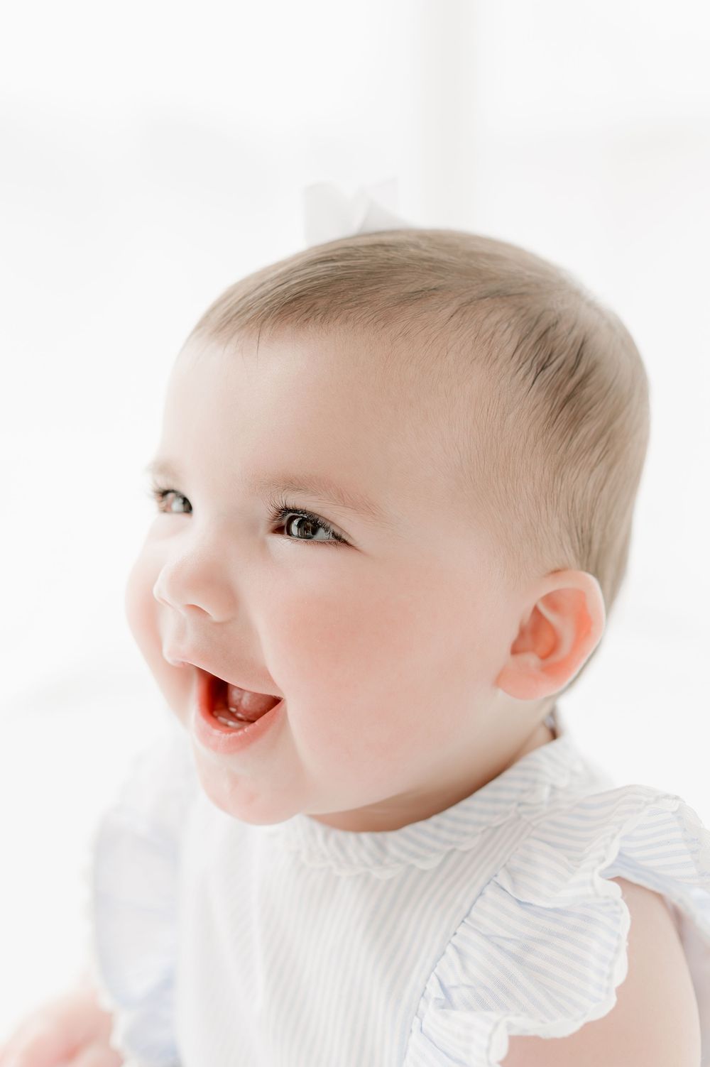 A happy baby in white clothing smiles and laughs against a bright white background.