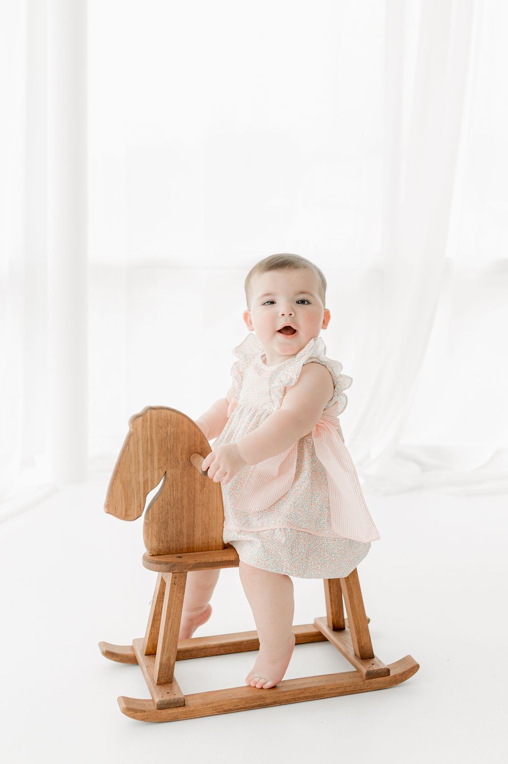 Baby in white dress plays on wooden rocking horse against bright white background in studio setting.
