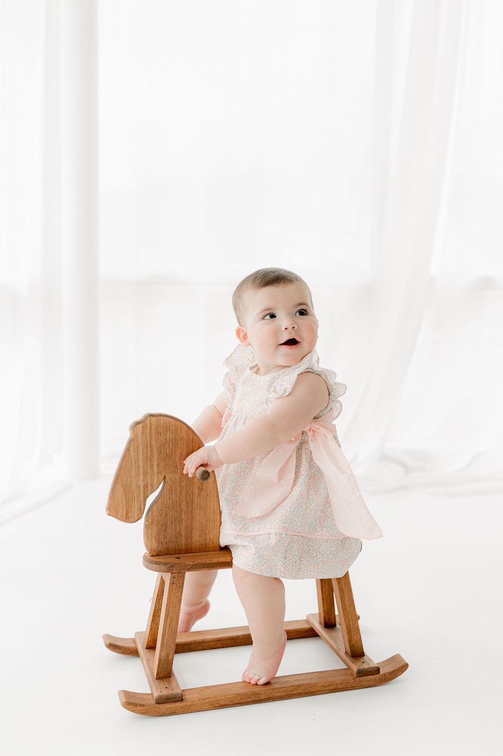 A baby in a white dress sits and plays on a wooden rocking horse toy in a bright white room.