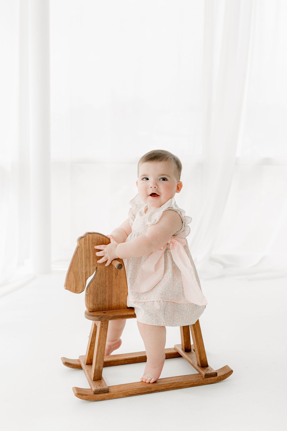 Baby in white dress stands on wooden rocking horse toy against bright white background.