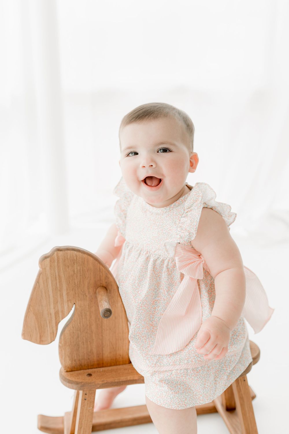 Baby in white lace dress smiles while sitting on wooden rocking horse against bright background.
