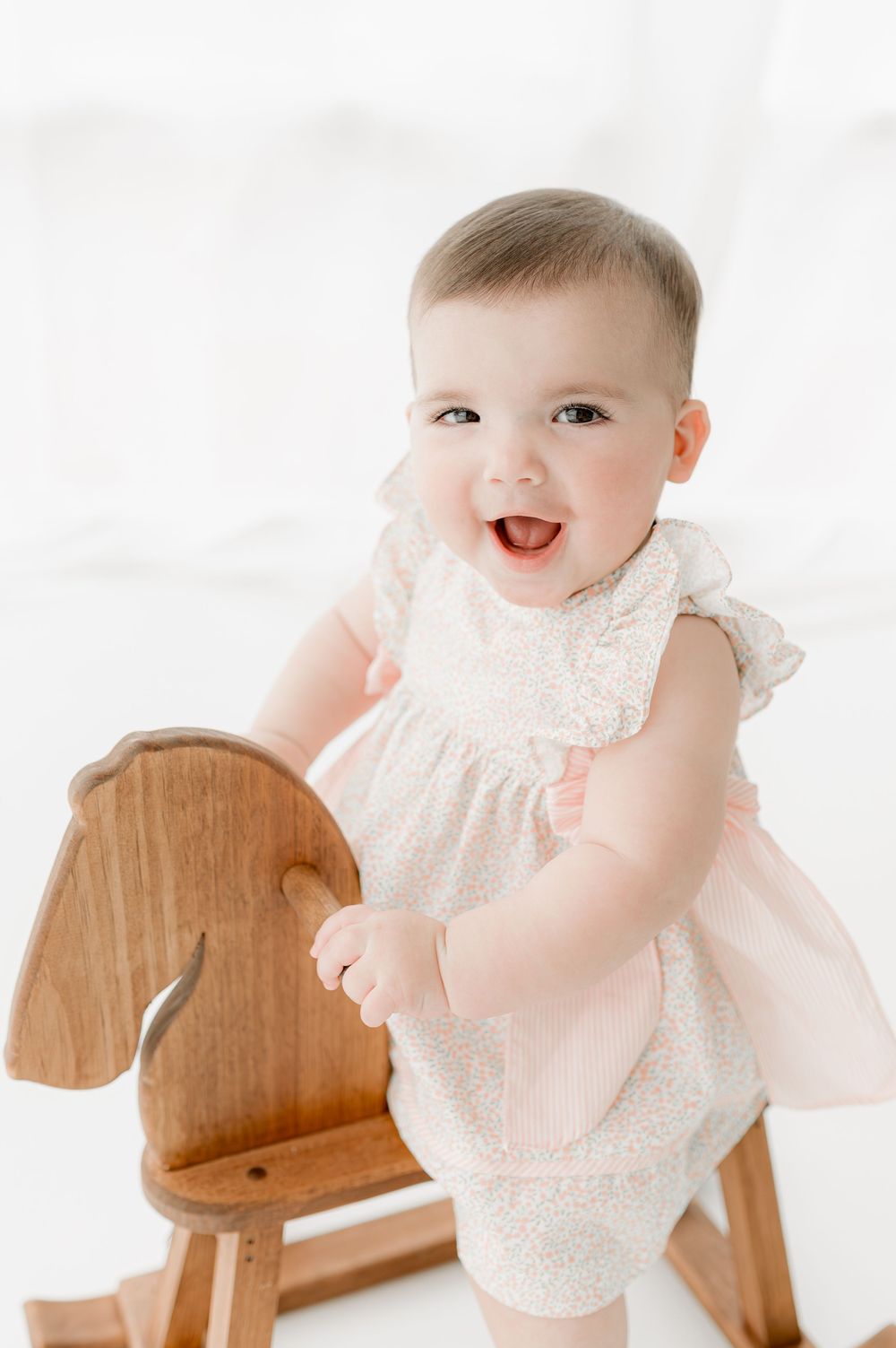 Happy baby in white lace dress smiles while sitting on wooden rocking horse in bright studio setting.
