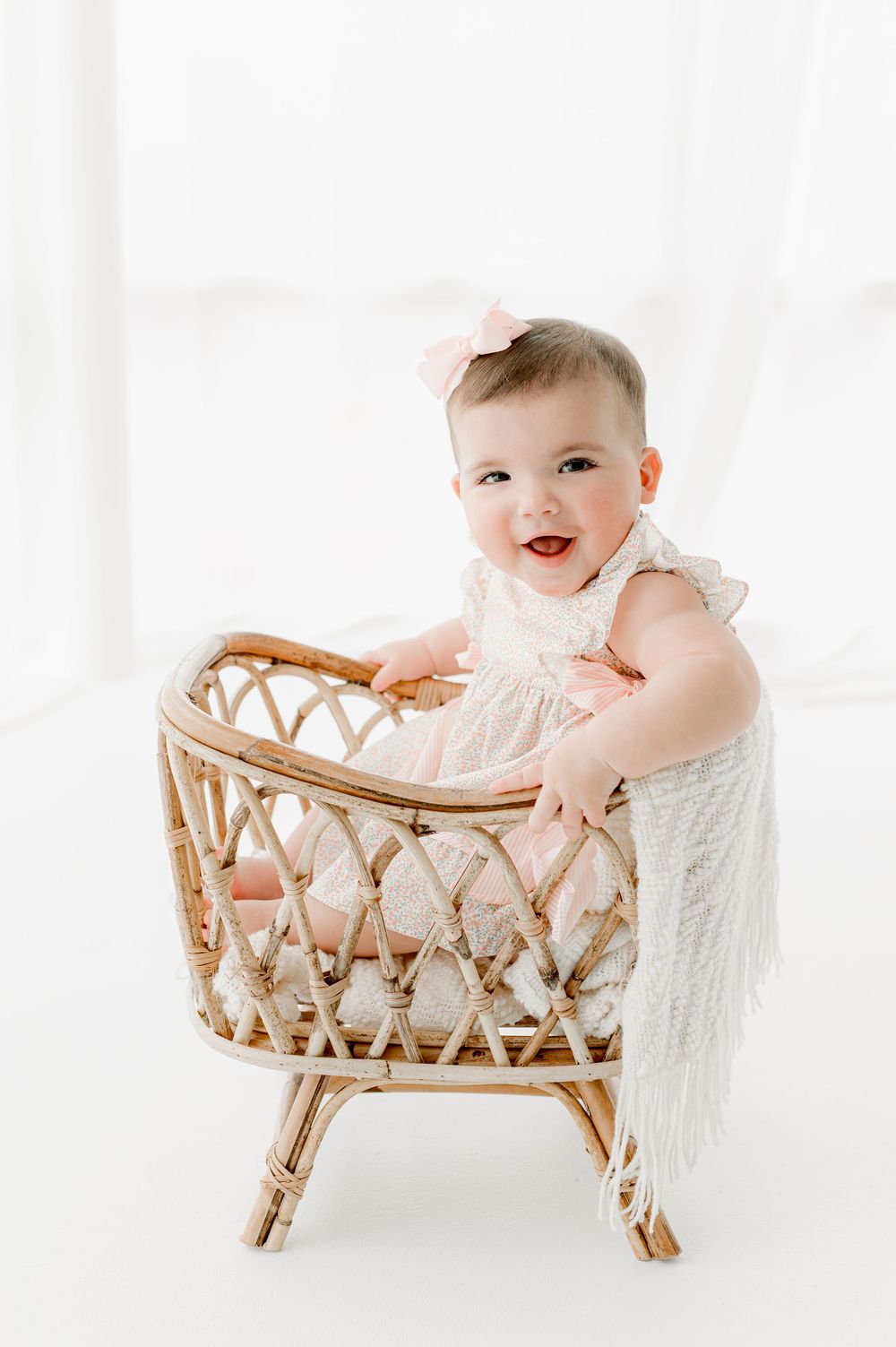 A baby lies in a woven rattan bassinet wearing a white outfit and smiling joyfully against a bright white background.