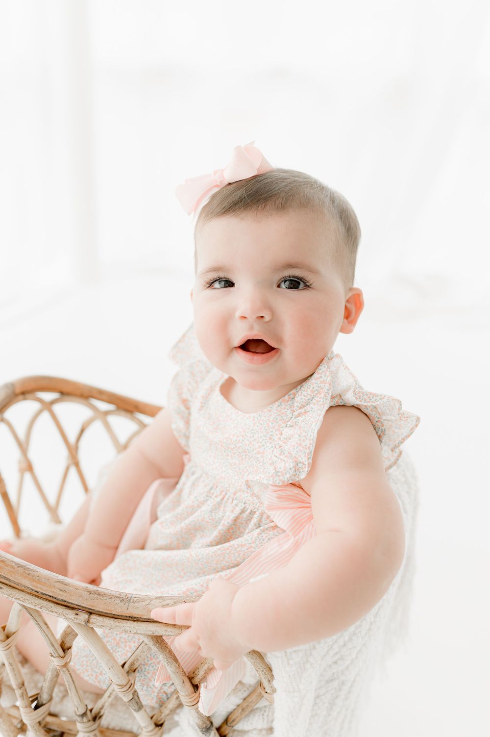 A baby in a white lace dress sits in a vintage wicker chair against a bright white background.