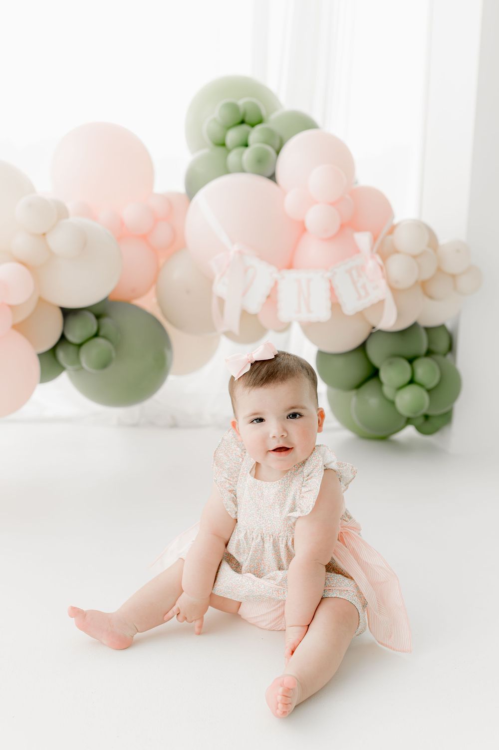 Baby in white dress sitting on floor with pink and sage green balloon arch decorations in bright studio setting.