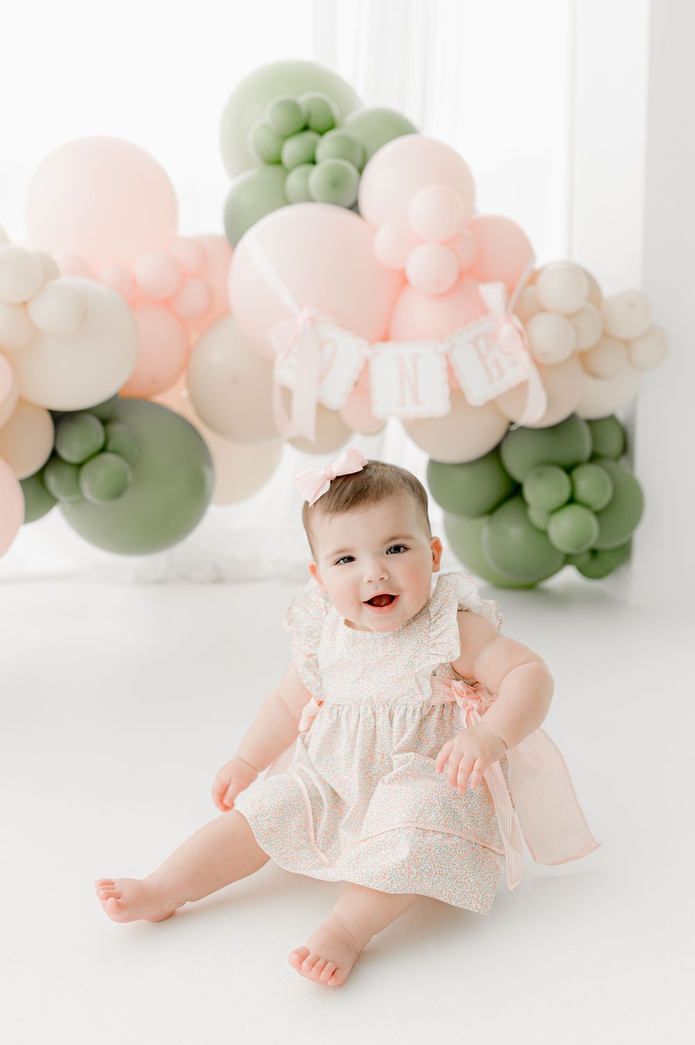 Sweet baby in pink dress sits among soft pastel balloon decorations in green and peach colors for a photo session.