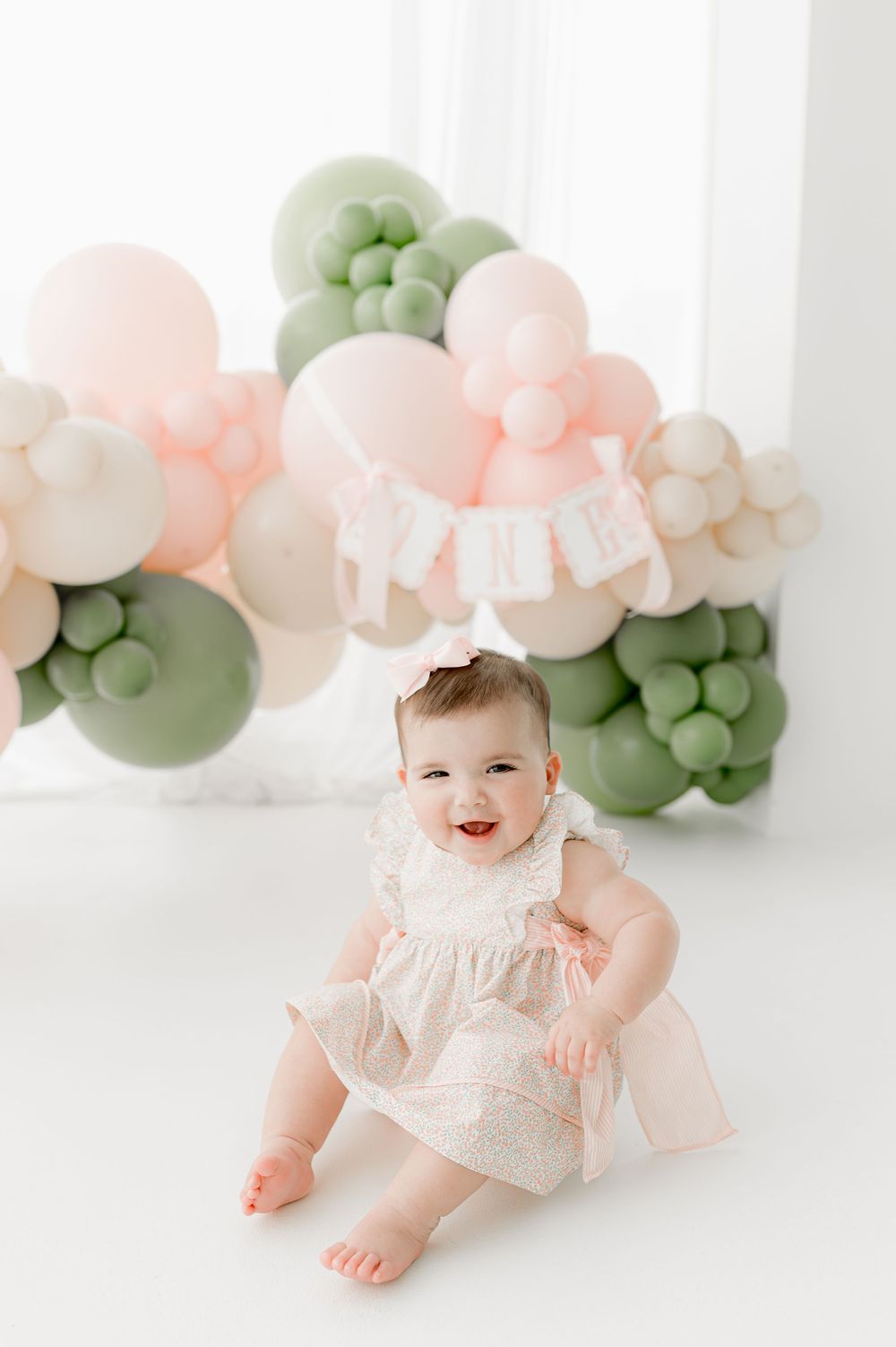 A baby in a white lace dress sits against a backdrop of soft pink and sage green balloon decorations.