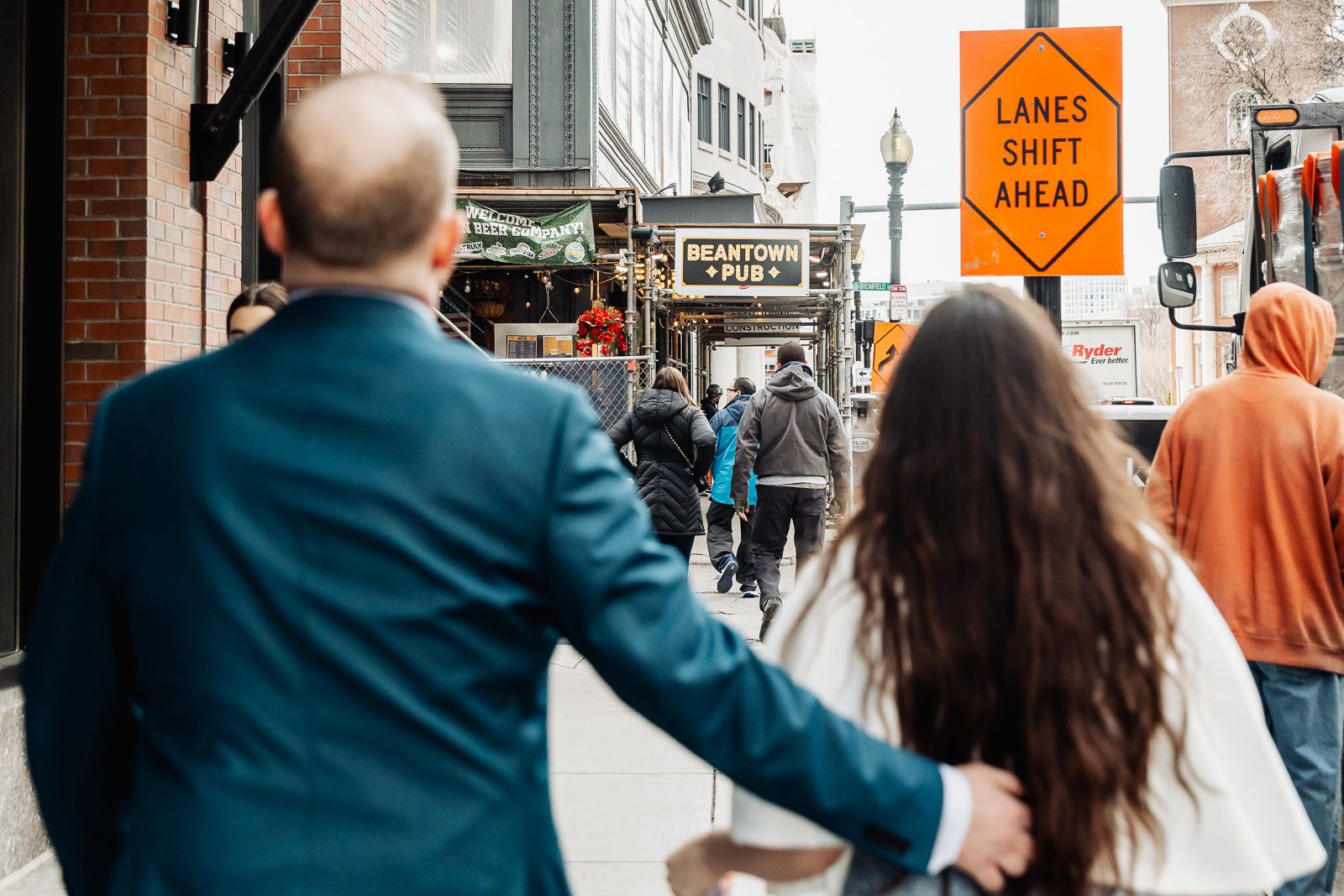 Couple walking hand-in-hand on a busy city sidewalk past an orange lanes shift ahead construction sign.