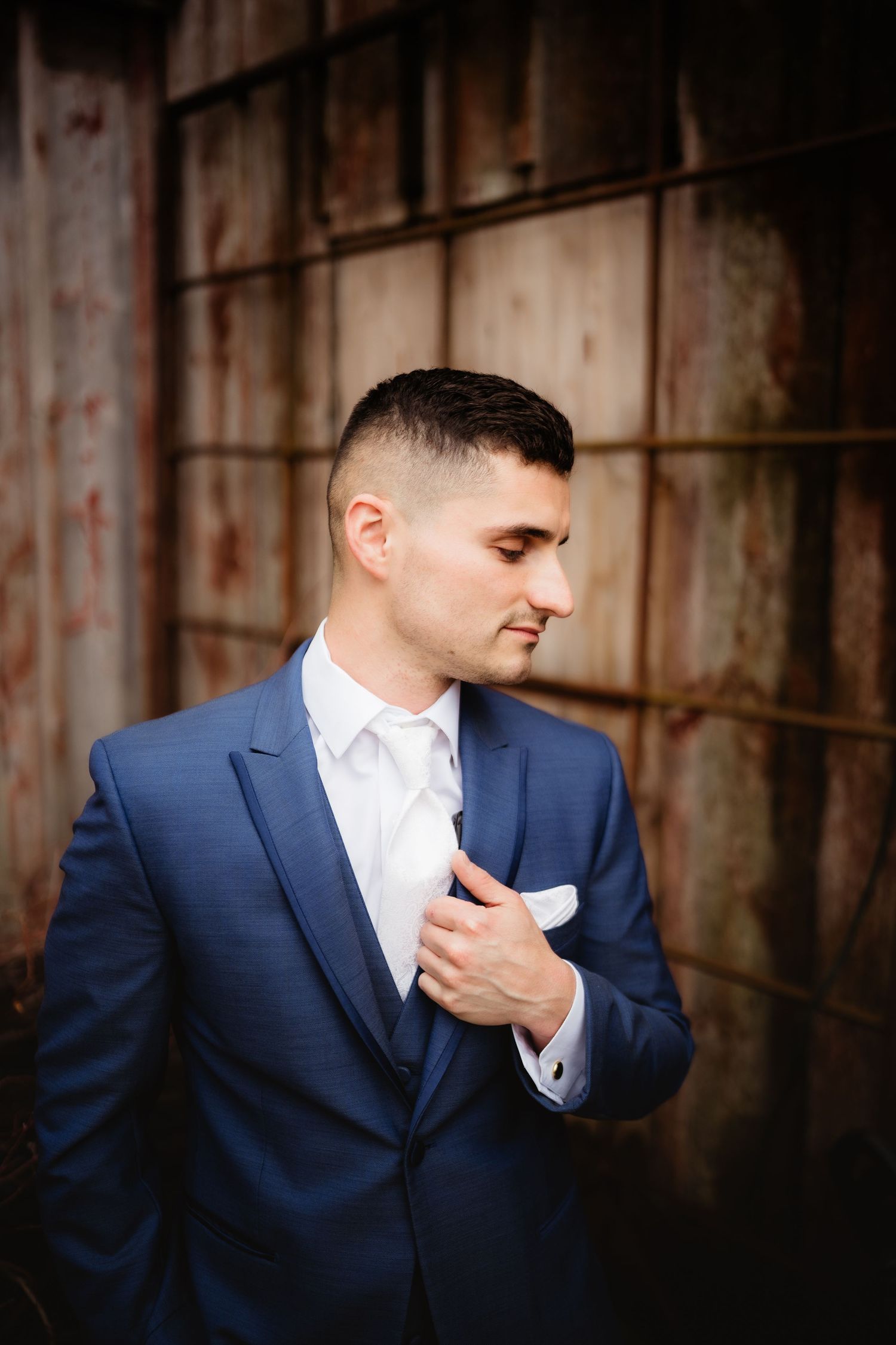 A well-dressed groom in a navy blue suit adjusts his white tie and pocket square against a rustic wooden backdrop.