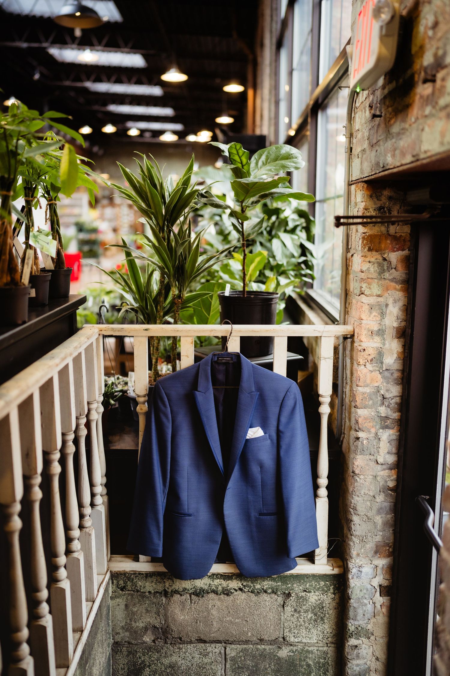 Navy blue suit jacket with white pocket square hanging on staircase railing surrounded by lush green plants in rustic venue.