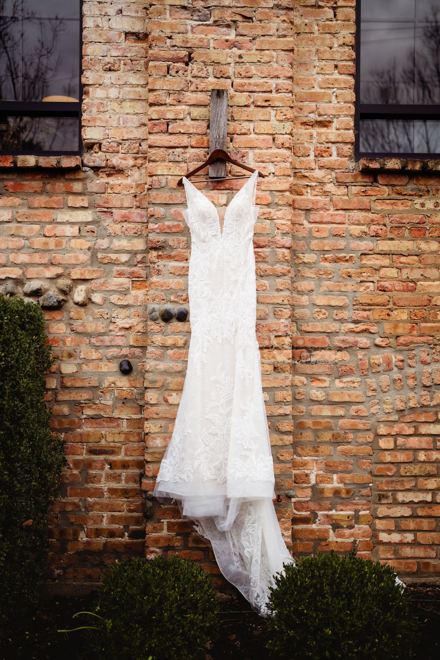 White lace wedding dress hanging on wooden hanger against rustic brick wall outdoors.