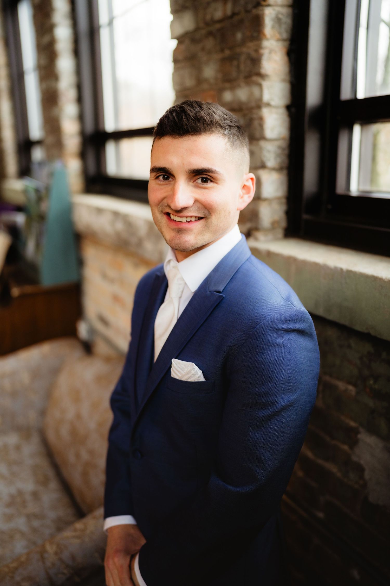 Smiling young man in a navy blue suit with white pocket square poses indoors near a brick wall and window.