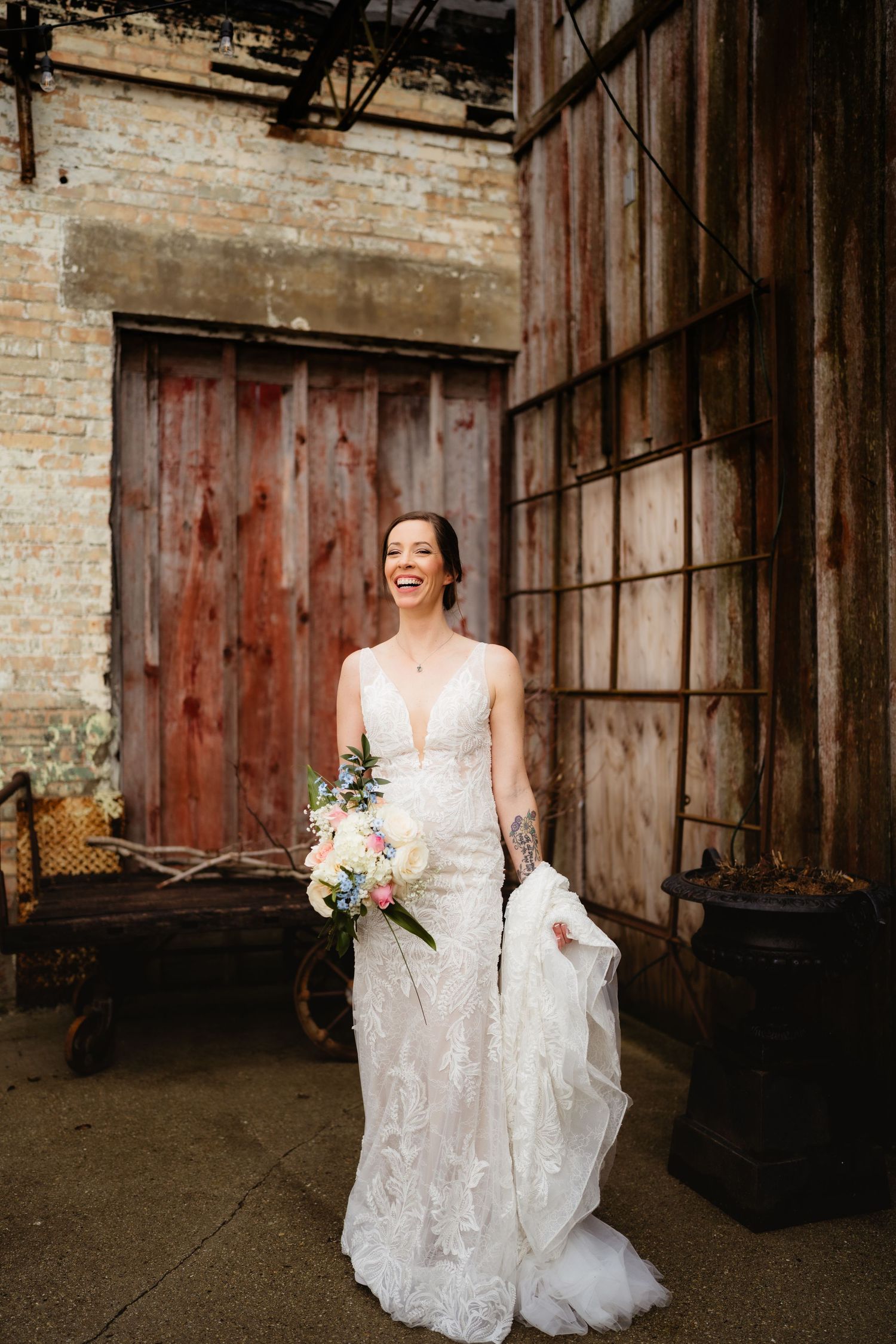 Laughing bride in deep V-neck lace wedding gown holding bouquet outside rustic industrial building with weathered wood doors.