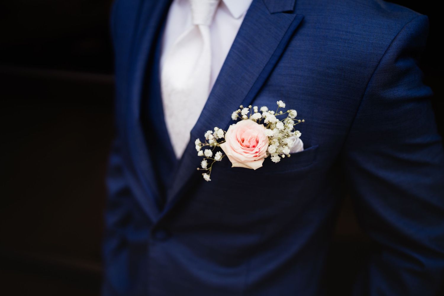 Close-up of a groom in a navy blue suit wearing a pink rose and baby's breath boutonniere on his lapel.