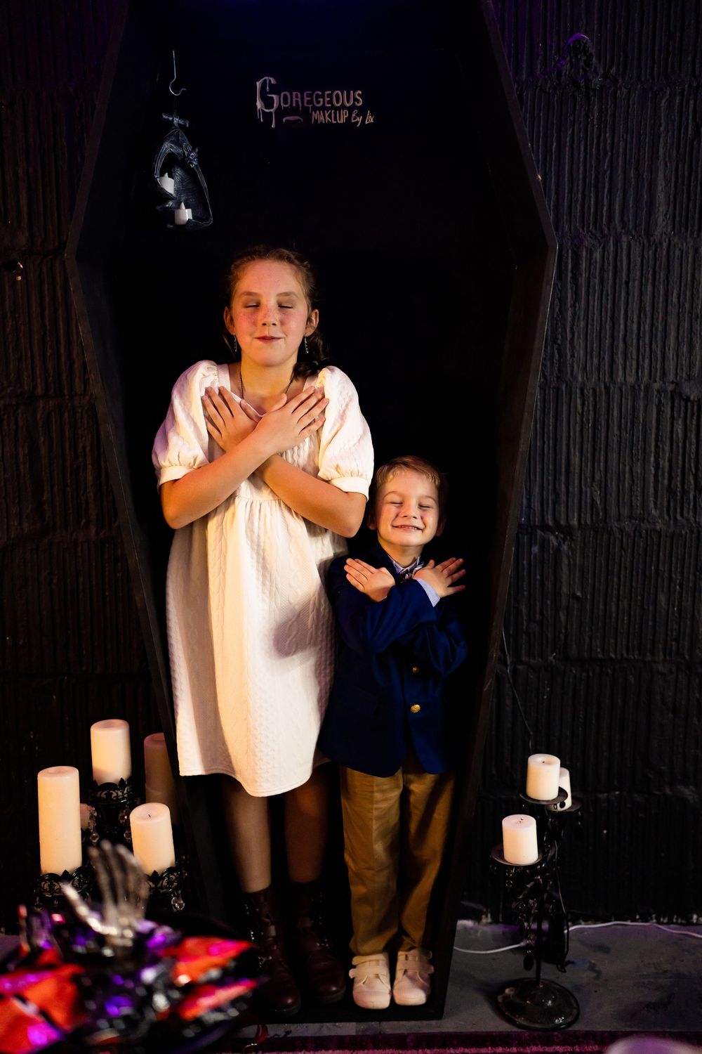 Two children in Halloween costumes pose inside a spooky coffin prop with candelabras and skeleton decorations.