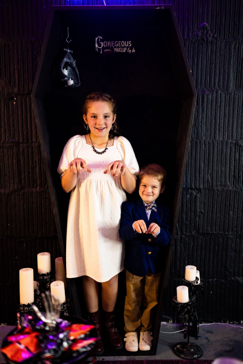 Two children pose in a spooky Halloween coffin backdrop with candelabras for a festive photo shoot.