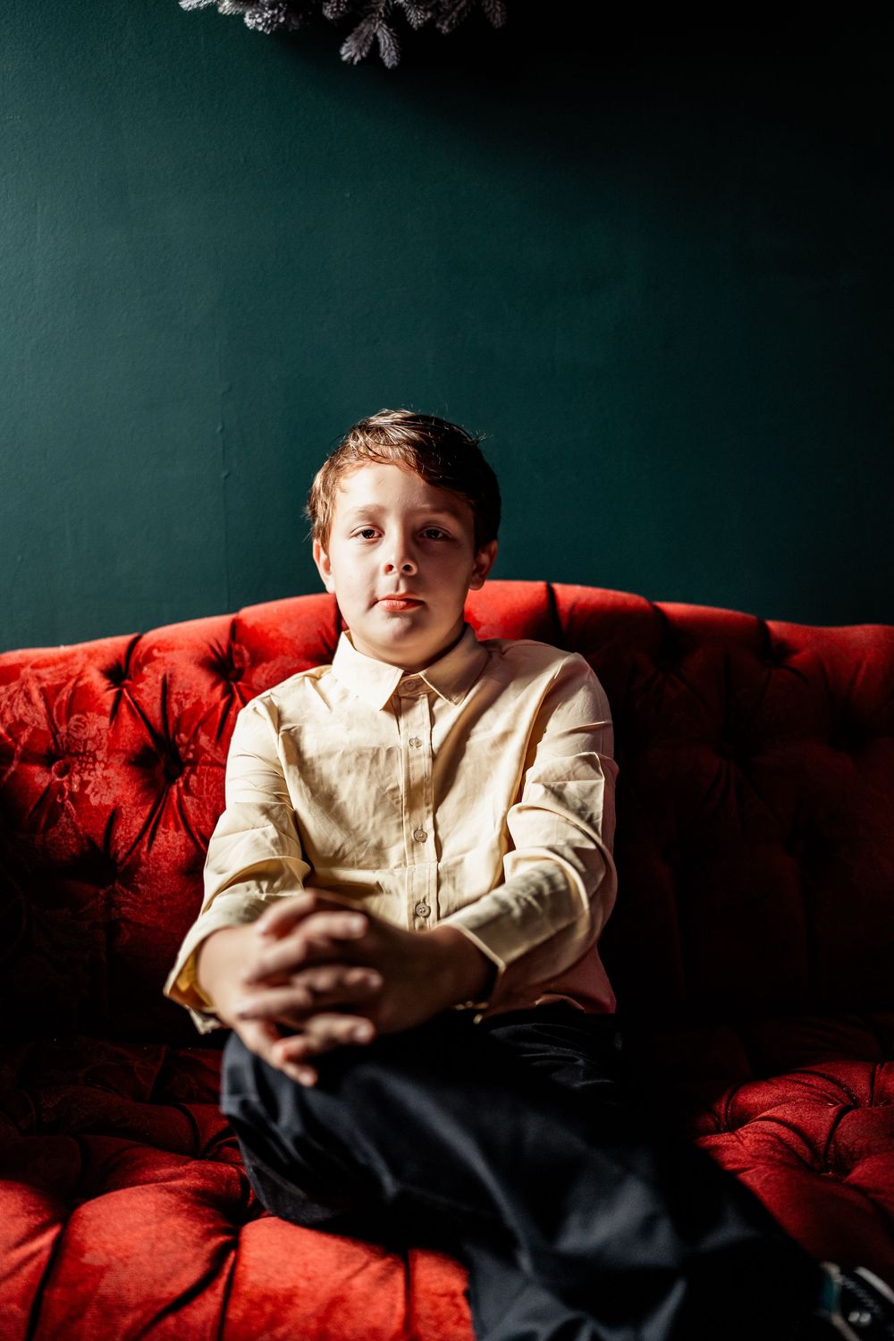 Boy in cream shirt sits on red tufted velvet sofa against dark green wall with holiday decor.