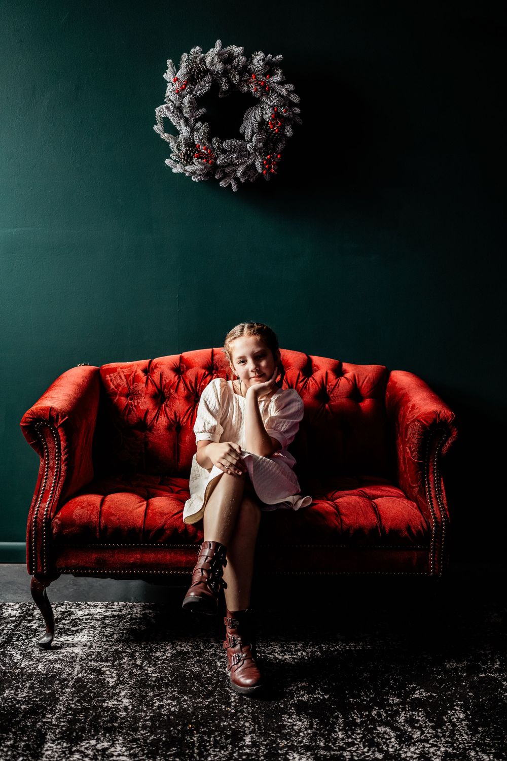 Girl in white dress and boots sits on red velvet sofa with Christmas wreath on dark green wall.