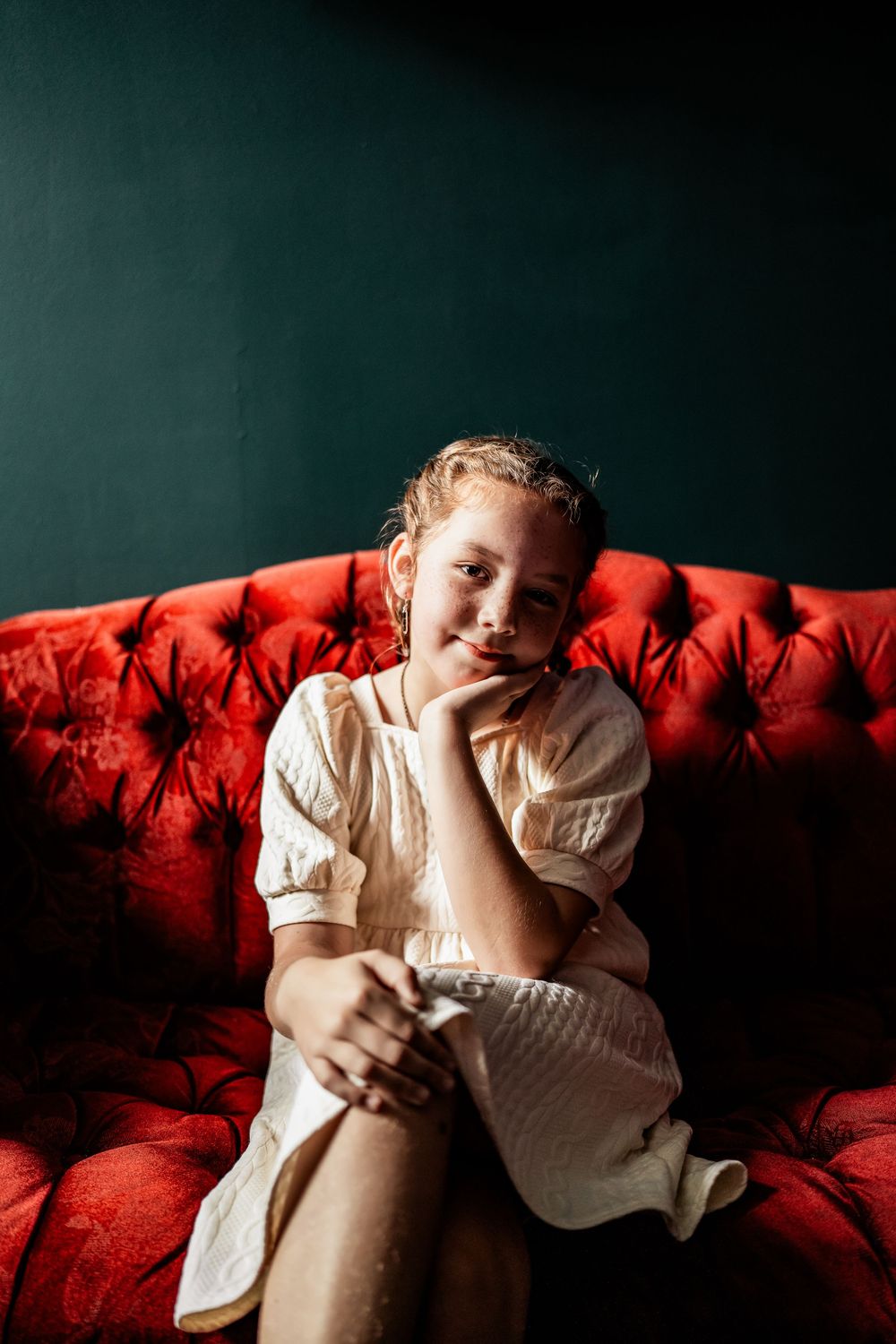 Young girl in white dress sitting thoughtfully on red tufted velvet sofa against dark green wall.