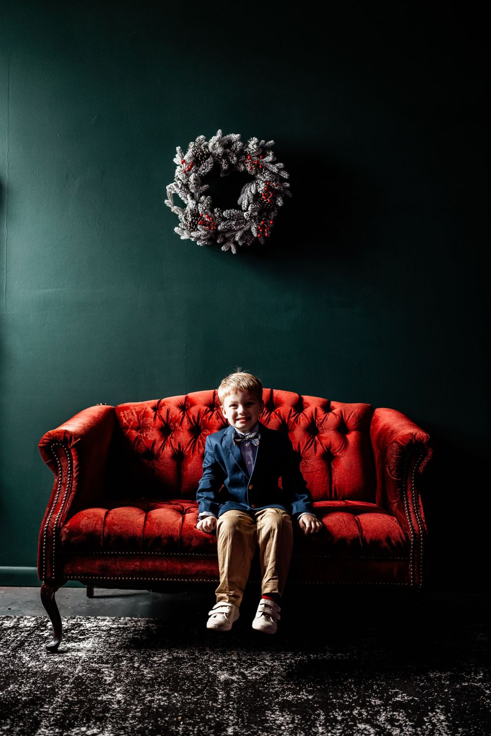 Young boy in navy blazer sitting on red velvet sofa with Christmas wreath on dark green wall.