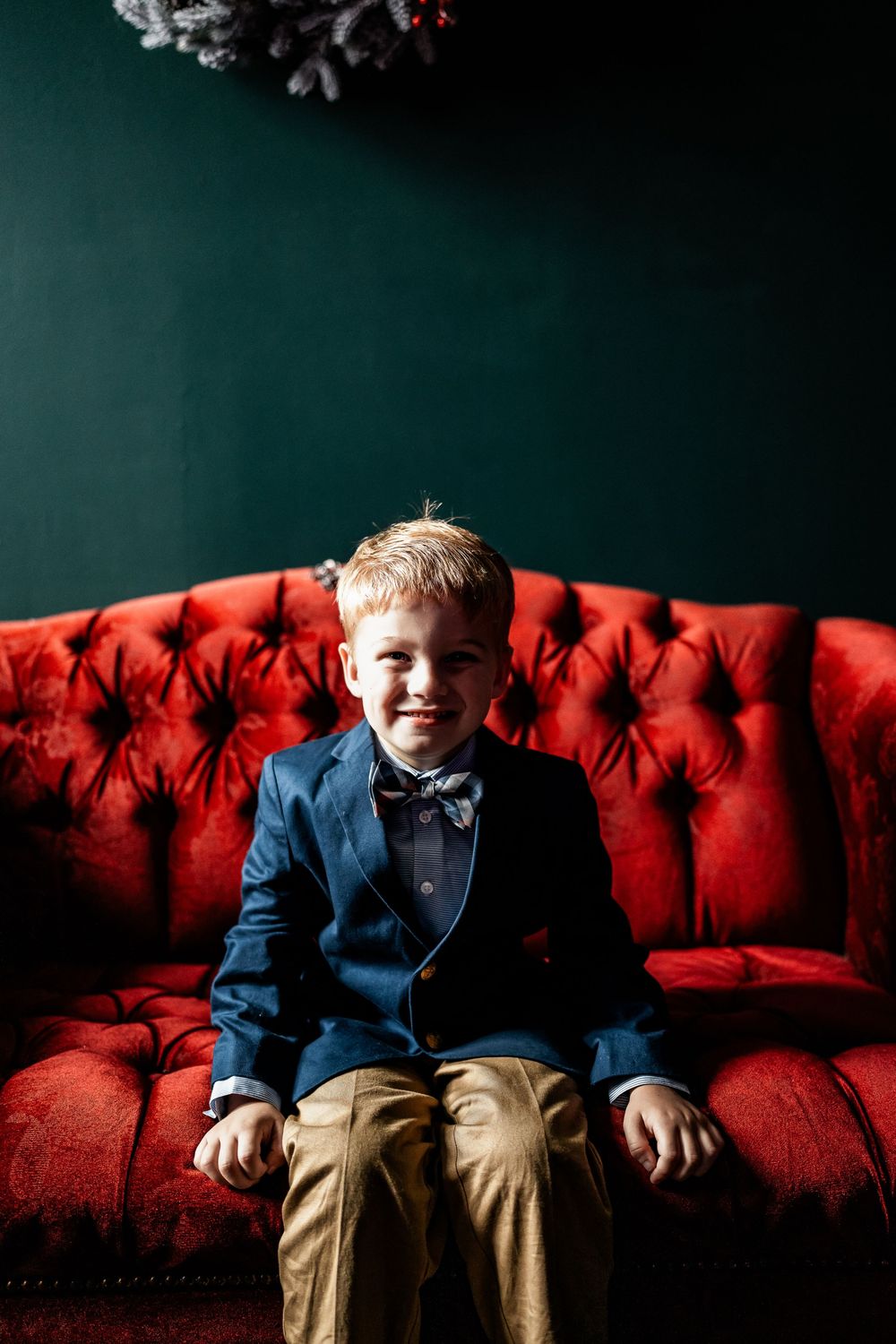 Young boy in navy blazer and bow tie smiling on red tufted velvet sofa with green wall backdrop.
