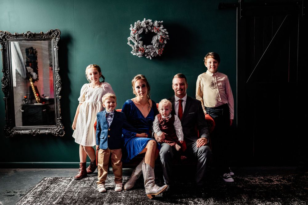 Family of six poses for a festive Christmas portrait against a dark teal wall with holiday wreath decor.