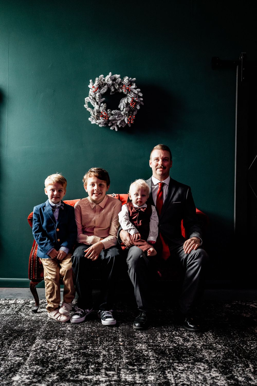 A father and three young sons pose for a Christmas family portrait on a red sofa with holiday wreath.