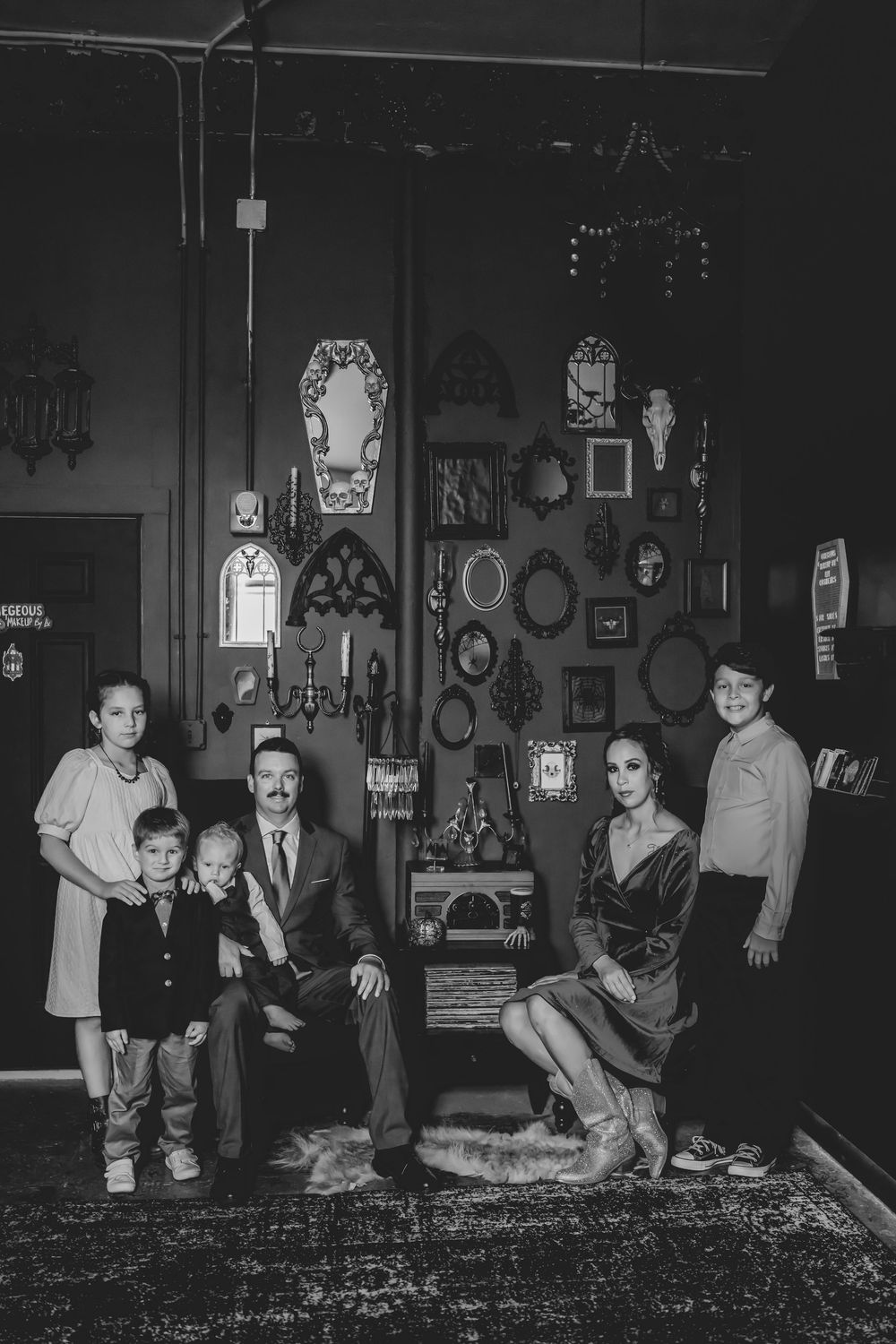 Black and white family portrait of six people posed in vintage room with gallery wall of ornate frames.