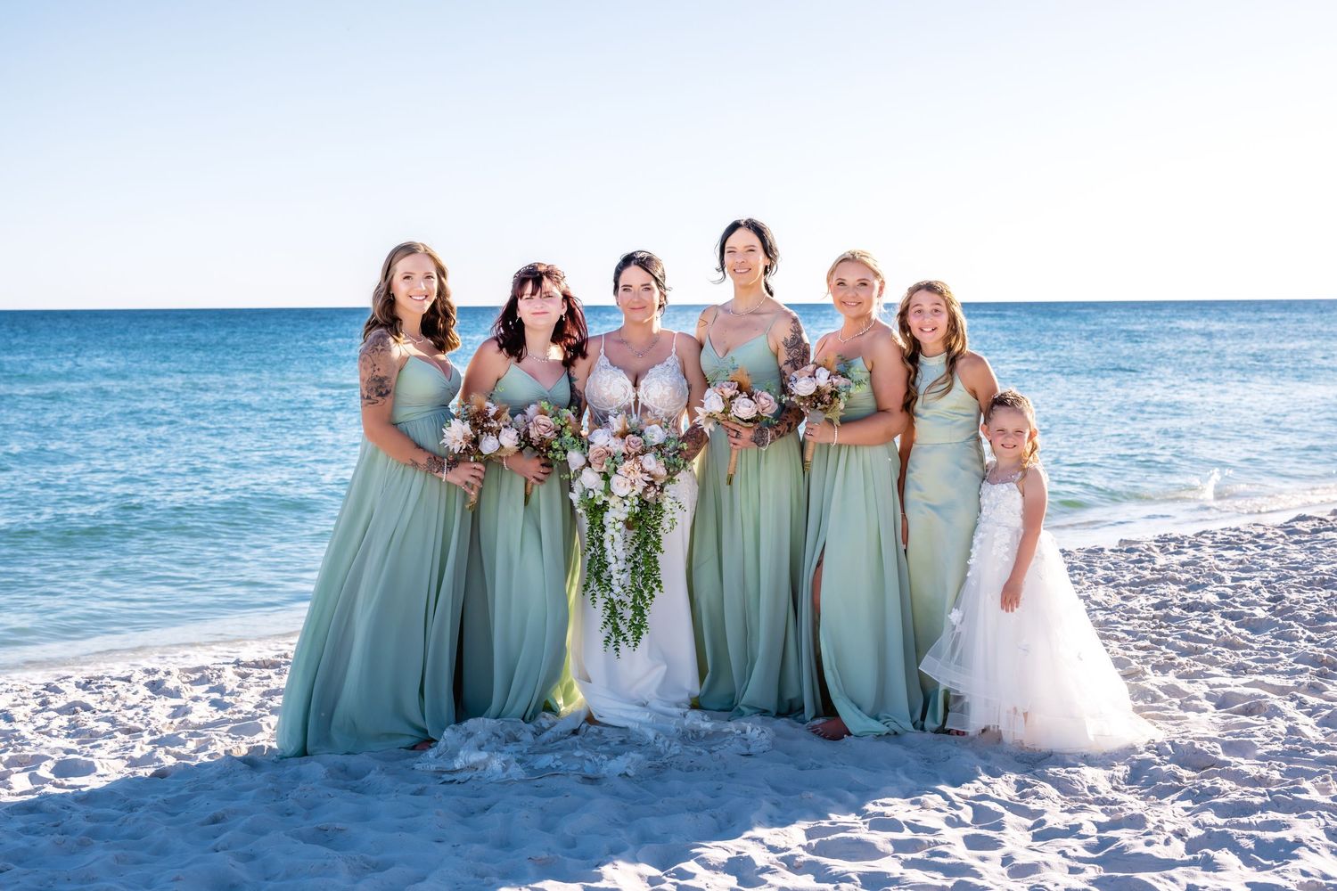 Bride and bridesmaids in sage green gowns pose on a sunny beach with ocean backdrop holding pink bouquets.